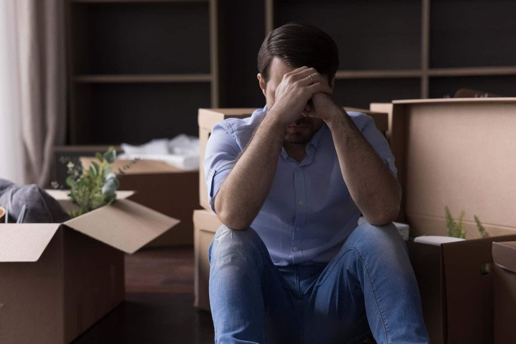 istockphoto-1485486081-1024×1024 Gestresster Mann in Jeans und Hemd sitzt vor Umzugskartons und hält den Kopf in den Händen.