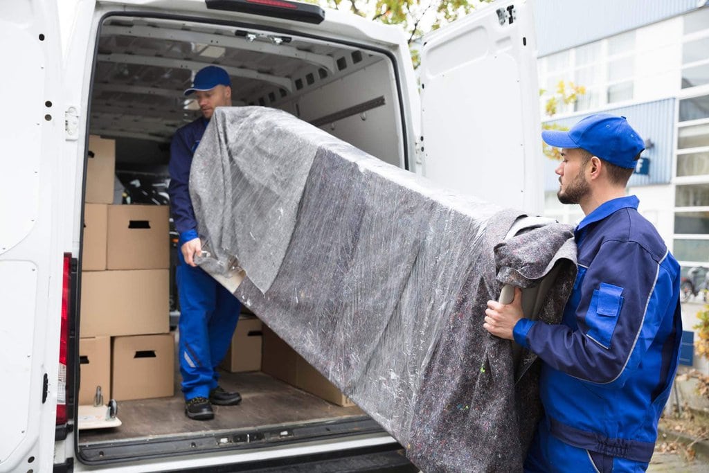 Umzugshelfer tragen ein verpacktes Möbelstück aus einem Transporter mit Kartons im Hintergrund