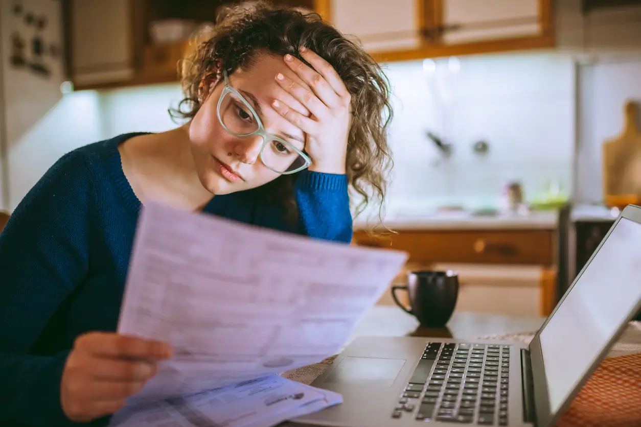 iStock-1013076770 Eine Frau sieht gestresst aus, während sie in einer Küche an ihrem Laptop Papiere durchgeht.