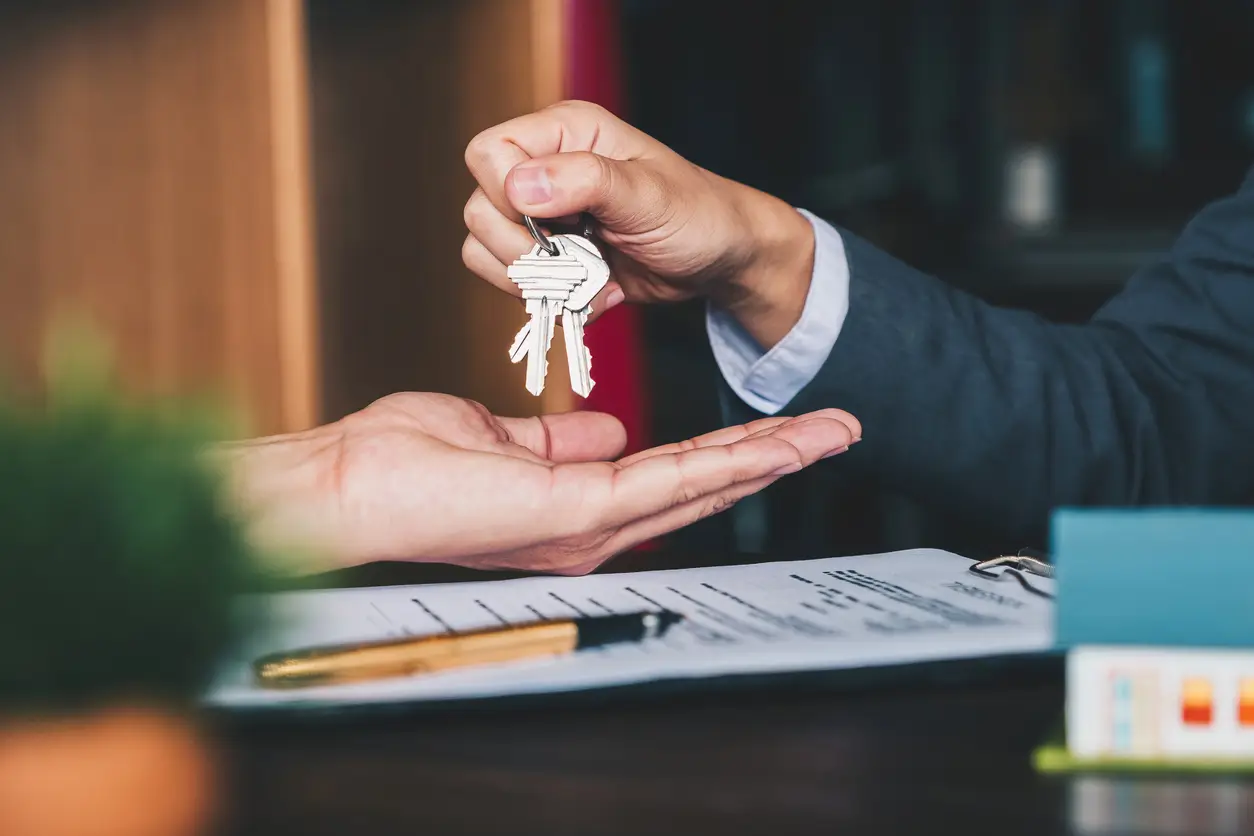 iStock-1130829500 Eine Person übergibt einer anderen die Hausschlüssel, während auf einem Schreibtisch ein Mietkautionsvertrag und ein Stift liegen.