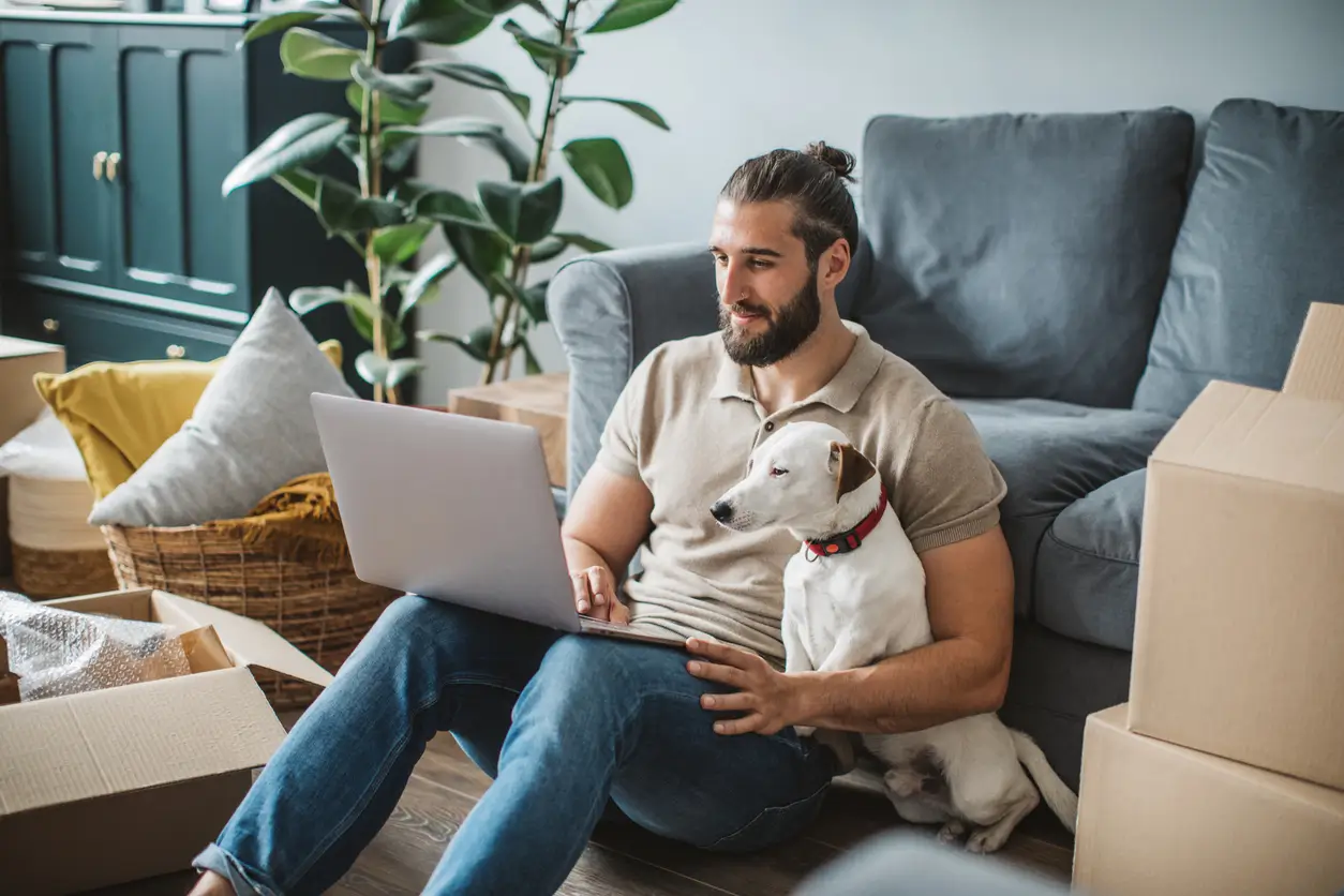 iStock-1313346962 Mann sitzt mit Laptop und Hund zwischen Umzugskartons – bereit zum Umzug melden Zürich.