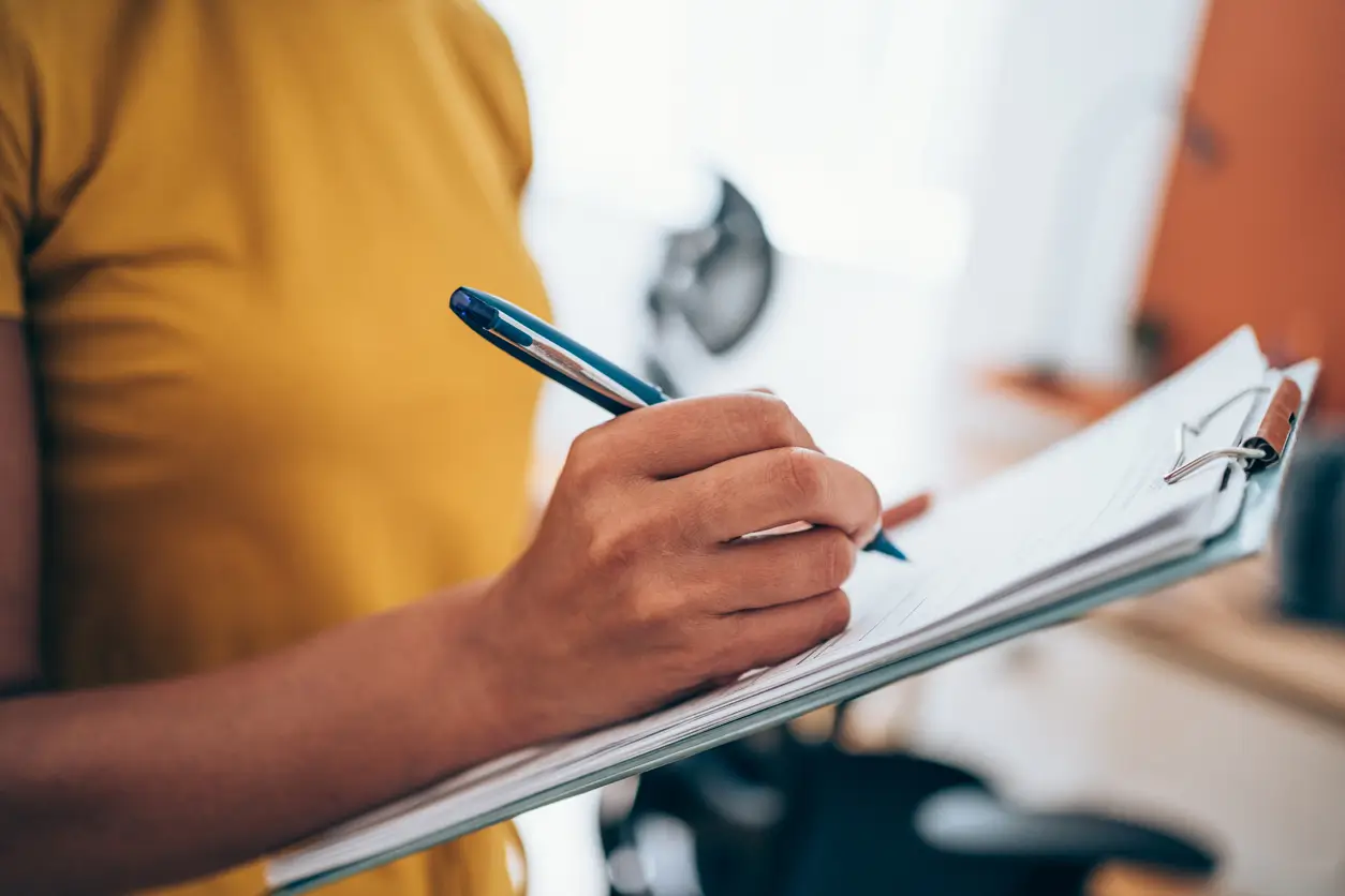 Eine Person in einem gelben Hemd schreibt mit einem blauen Stift eine Umzugs-Checkliste auf ein Klemmbrett.